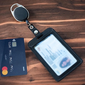 May include: A black leather ID badge holder with a clear plastic window and a retractable reel clip. The holder is stitched with black thread and features a silver metal snap button. A dark blue debit card with a contactless symbol is placed nearby.