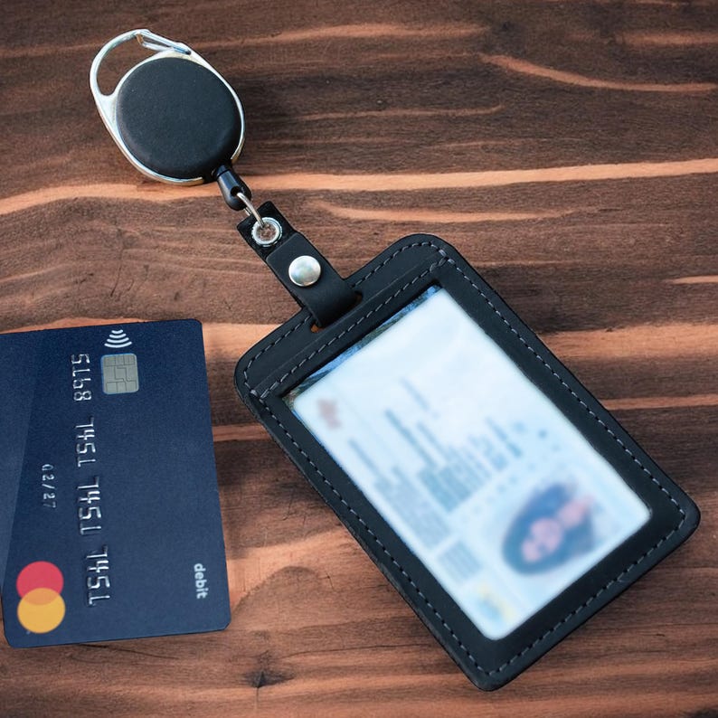 May include: A black leather ID badge holder with a clear plastic window and a retractable reel clip. The holder is stitched with black thread and features a silver metal snap button. A dark blue debit card with a contactless symbol is placed nearby.