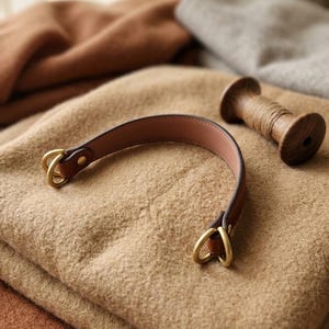 Puede incluir: Una correa de cuero marrón curvada con anillos de metal dorado en cada extremo, colocada sobre una tela de lana beige doblada junto a un pequeño carrete de madera envuelto con hilo marrón, con más telas dobladas al fondo.