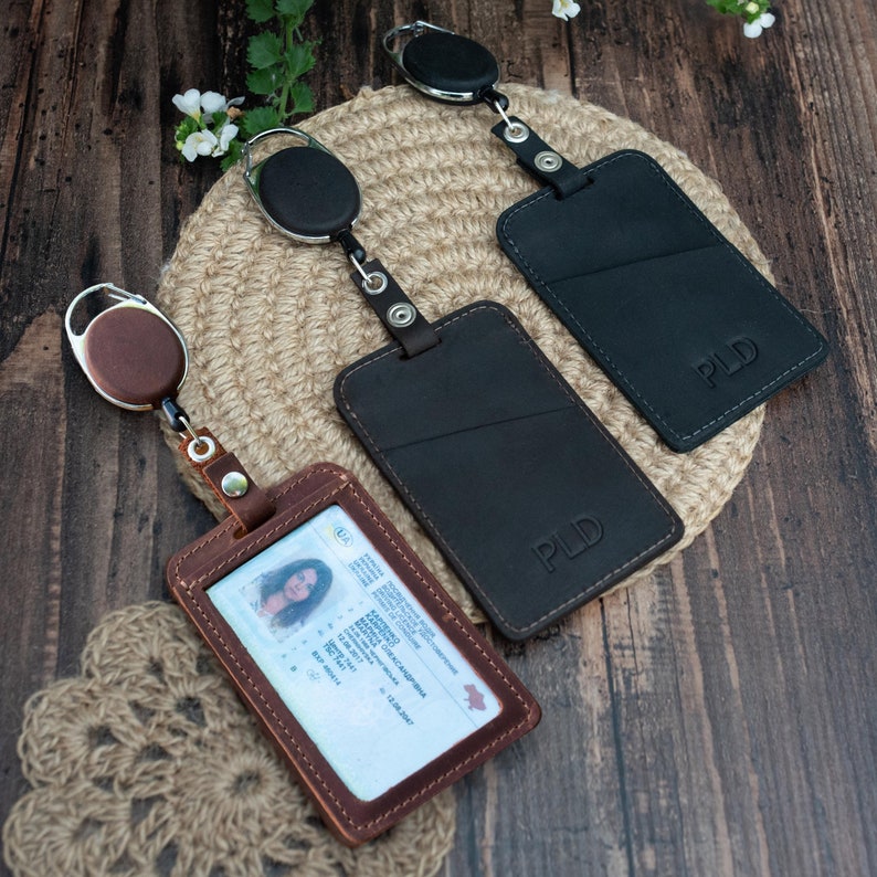 May include: Three rectangular badge holders with retractable reels. One brown holder displays a driver's license with text, while two black holders are embossed with the letters PLD. Each holder features a clear window and a metal snap closure.