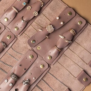 May include: A mauve leather jewelry organizer roll featuring multiple snap-button tabs for securing rings and earrings, along with integrated slots for necklaces. The interior is lined with soft material and holds various gold and silver rings.