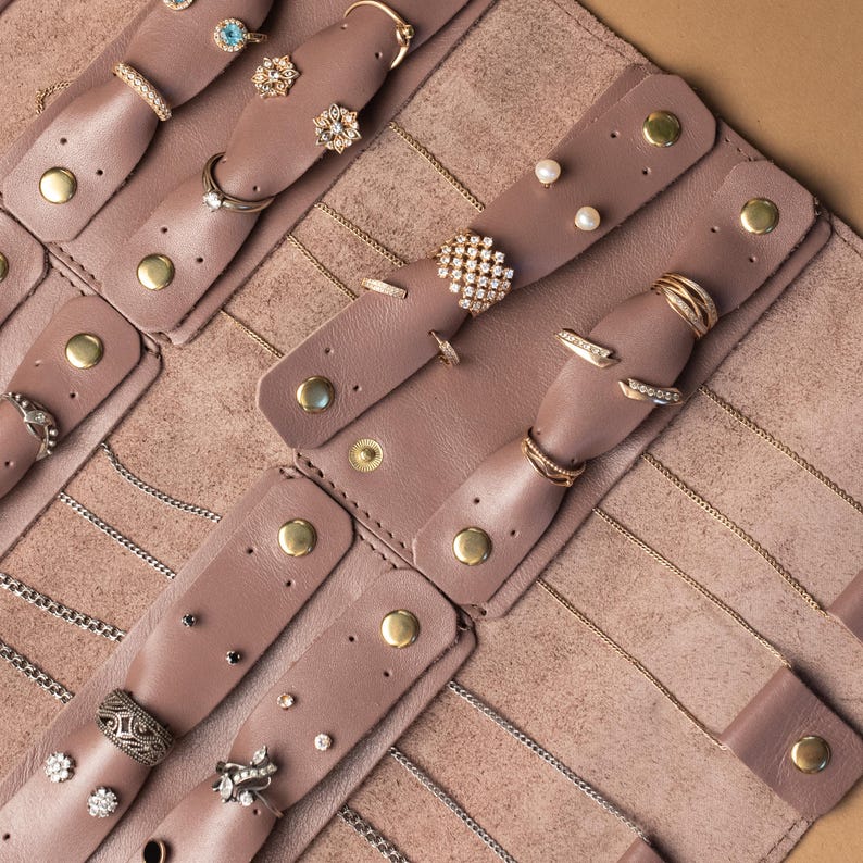 May include: A mauve leather jewelry organizer roll featuring multiple snap-button tabs for securing rings and earrings, along with integrated slots for necklaces. The interior is lined with soft material and holds various gold and silver rings.