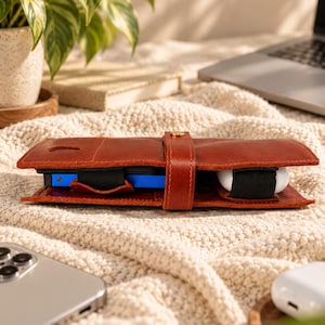 Peut inclure: Un étui organisateur en cuir marron doté d'une sangle centrale avec une fermeture à bouton-pression en métal doré. L'intérieur contient un appareil électronique rectangulaire bleu et un boîtier d'écouteurs sans fil blanc, fixés par des élastiques noirs.