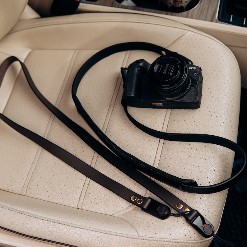 Puede incluir: Una c&aacute;mara Canon EOS R negra con una tapa de lente y una correa de c&aacute;mara de cuero negro. La correa tiene remaches dorados y detalles en rojo. La c&aacute;mara y la correa est&aacute;n sobre un asiento de coche de cuero crema perforado.