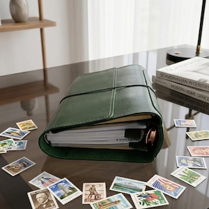May include: A dark green leather-bound planner with a black elastic closure band, containing white paper pages and a metal ring binder mechanism, surrounded by a collection of various vintage postage stamps on a dark reflective surface.