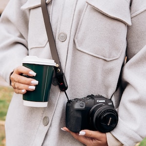 Puede incluir: Una c&aacute;mara Canon EOS negra con una lente es sostenida por una persona. La c&aacute;mara est&aacute; sujeta a una correa marr&oacute;n. Una taza de caf&eacute; verde oscuro con tapa blanca se sostiene en la otra mano. La persona lleva una chaqueta gris claro.
