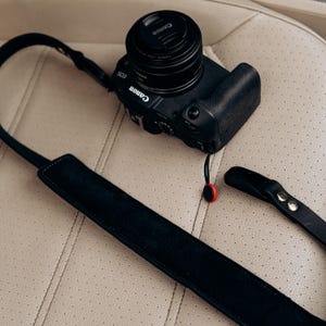 Puede incluir: Una c&aacute;mara Canon EOS negra con tapa de lente, sobre una superficie beige claro. Una correa de c&aacute;mara negra est&aacute; adjunta, con una almohadilla para el hombro de gamuza. La correa est&aacute; parcialmente separada, mostrando un conector rojo y negro.