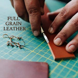 May include: A strip of brown full grain leather being cut with a metal craft knife on a green cutting mat with yellow grid lines. Small curled leather shavings lie on the mat. The text FULL GRAIN LEATHER is printed in brown on the left side.