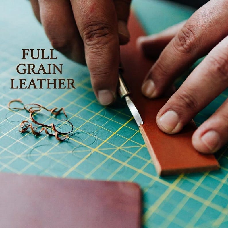 May include: A strip of brown full grain leather being cut with a metal craft knife on a green cutting mat with yellow grid lines. Small curled leather shavings lie on the mat. The text FULL GRAIN LEATHER is printed in brown on the left side.