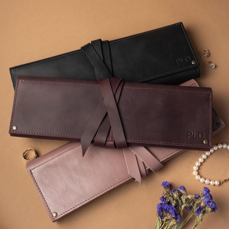 May include: Three rectangular leather cases in black, dark burgundy, and dusty rose, each featuring a wrap-around tie closure, visible stitching along the edges, metallic rivets at the corners, and the embossed letters PLD on the bottom right corner.