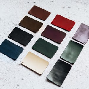 May include: A collection of eleven rectangular leather swatches arranged in a grid pattern. The swatches feature a variety of colors including brown, red, black, dark green, navy blue, tan, and purple, each with a small circular hole in one corner.