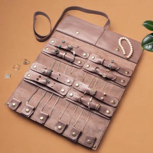 May include: A mauve leather jewelry roll organizer featuring multiple snap-button straps for securing necklaces, rings, and earrings. The interior includes dedicated sections for various accessories, with a top flap and a strap for closure.