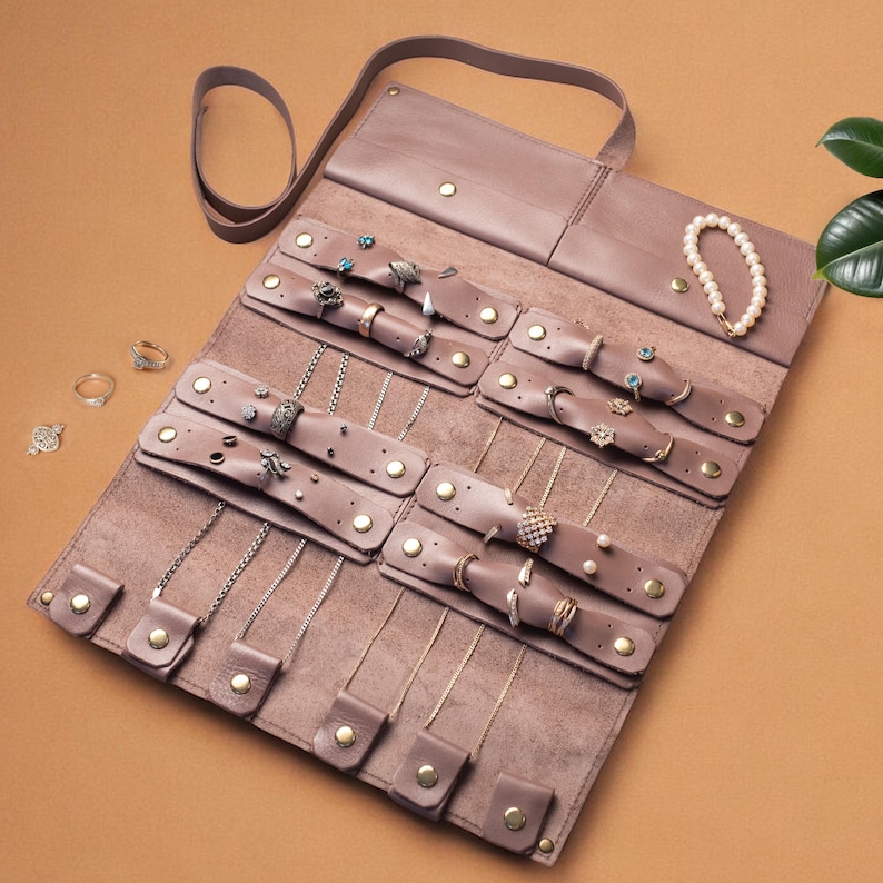 May include: A mauve leather jewelry roll organizer featuring multiple snap-button straps for securing necklaces, rings, and earrings. The interior includes dedicated sections for various accessories, with a top flap and a strap for closure.