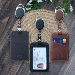 May include: Three rectangular badge holders with retractable carabiner reels. Two are dark brown with the embossed letters PLD, and one is black with a clear plastic window displaying an identification card. Each holder features a vertical slot.