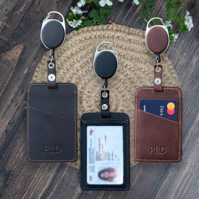 May include: Three rectangular badge holders with retractable carabiner reels. Two are dark brown with the embossed letters PLD, and one is black with a clear plastic window displaying an identification card. Each holder features a vertical slot.