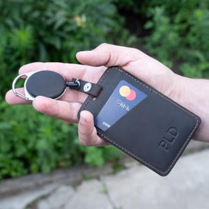 May include: A dark brown leather card holder with a retractable black badge reel attached by a metal snap. The card holder features a diagonal slot containing a blue payment card and is embossed with the letters PLD in the bottom right corner.