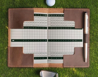 May include: A brown leather golf scorecard holder with stitched edges, containing a printed scorecard with numerical data. A silver pen is secured in a side loop, and a white dimpled golf ball and a metal golf club head are positioned nearby.