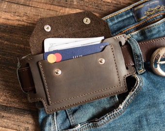 May include: A dark brown leather belt pouch featuring a flap with two silver-tone metal snap closures. The pouch is threaded onto a brown leather belt worn over blue denim jeans. A blue credit card and white paper currency are tucked inside the pouch.