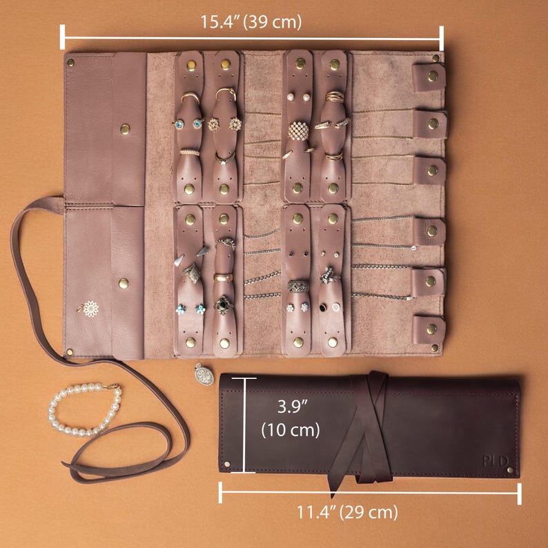 May include: A mauve leather jewelry roll organizer measuring 15.4 inches wide, featuring multiple snap-button tabs for necklaces and rings. A separate dark brown leather case measuring 11.4 inches by 3.9 inches is shown closed with a tie closure.