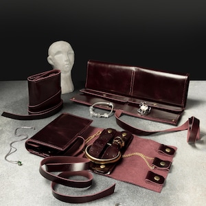 May include: A set of dark burgundy leather jewelry rolls with wrap-around straps and brass-colored snap closures. The rolls are displayed open and closed, containing various silver and gold-toned bracelets, rings, and necklaces, alongside a small white bust.