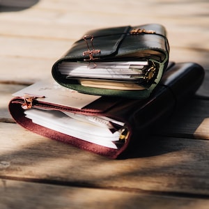 May include: Two leather journals stacked on top of each other. The top journal is dark green with a rose gold binder clip, and the bottom journal is deep burgundy with a matching rose gold binder clip. Both journals contain white paper inserts.