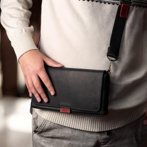 May include: Black crossbody bag with a flap closure and a red accent. The bag has a black strap with a red detail and a silver chain. The bag is rectangular and appears to be made of leather.