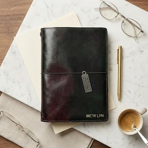 May include: A dark, multi-toned leather journal cover with a vertical elastic band and a rectangular metal charm engraved with the text I feel about you makes my heart lone to be free. The cover features the gold embossed initials MCW LPN.