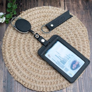 May include: A black leather badge holder with a clear window, attached to a retractable reel and a matching black leather keychain strap embossed with the letters PLD. The set features silver-toned metal hardware and visible stitching.