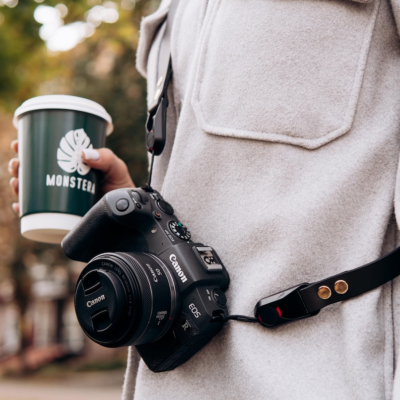 Puede incluir: Una c&aacute;mara Canon EOS R negra con tapa de lente y correa negra cuelga del cuello de una persona. La persona tambi&eacute;n sostiene una taza de caf&eacute; verde con la palabra "MONSTERA" impresa.