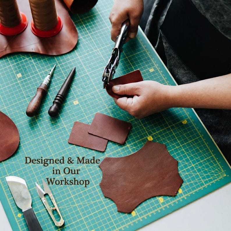 May include: A leather crafting scene featuring brown leather pieces, a metal hole punch tool, two wooden-handled awls, a metal blade, and metal snips on a green grid cutting mat. The text reads: Designed & Made in Our Workshop.