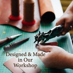 May include: A metal leather hole punch tool is used on a piece of brown leather. The text "Designed & Made in Our Workshop" is overlaid on the image. Other leatherworking tools and spools of thread are visible on a green cutting mat surface.