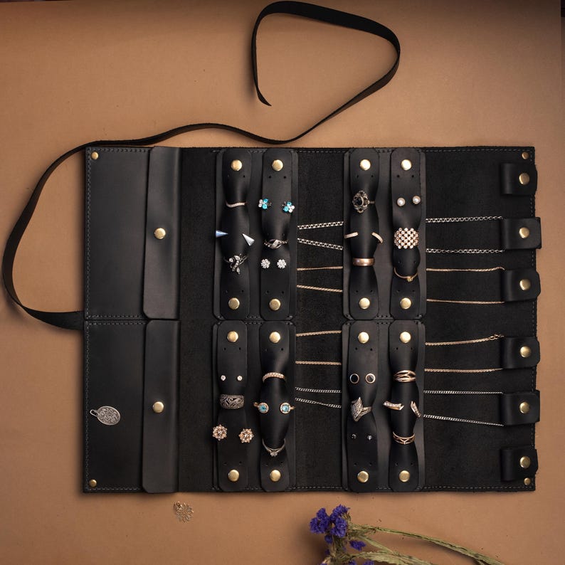 May include: A black leather jewelry roll organizer featuring multiple vertical straps with gold-tone snap buttons. The interior contains various rings, stud earrings, and delicate gold and silver chains secured by the leather tabs and snaps.
