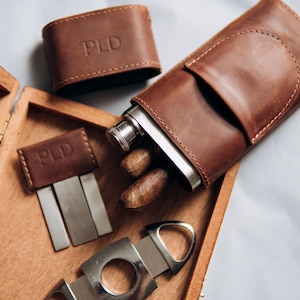 May include: A brown leather cigar case with a stainless steel flask and cigars. The case is embossed with the initials "PLD". A cigar cutter and a small leather case with cigar tools are also present.