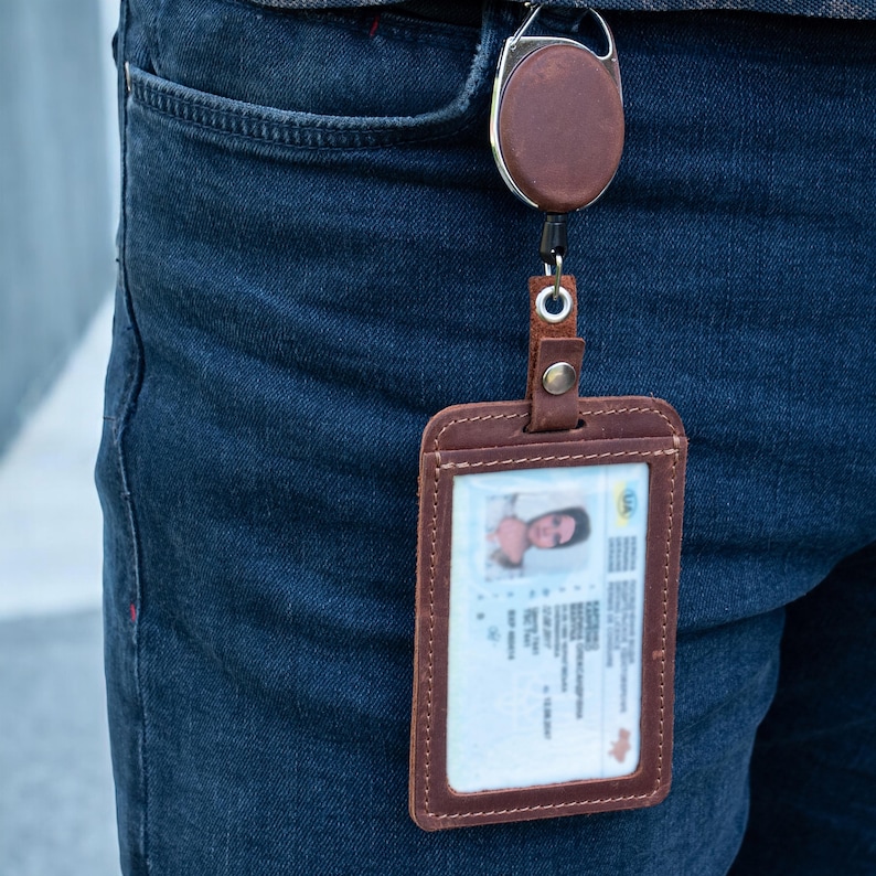 May include: A brown leather vertical ID badge holder with visible stitching, attached to a matching brown leather retractable reel clip. The holder contains a rectangular identification card with a photograph and printed text details.
