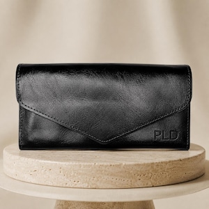 May include: A black rectangular wallet with a smooth, polished finish and an envelope-style flap closure. The bottom right corner features the embossed letters PLD. The wallet has visible stitching along the edges and a structured, slim profile.