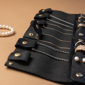 May include: A black leather jewelry roll organizer featuring multiple snap-button tabs to secure thin metal chains and a vertical strip with snap closures for rings and earrings, accompanied by a separate white pearl beaded bracelet.
