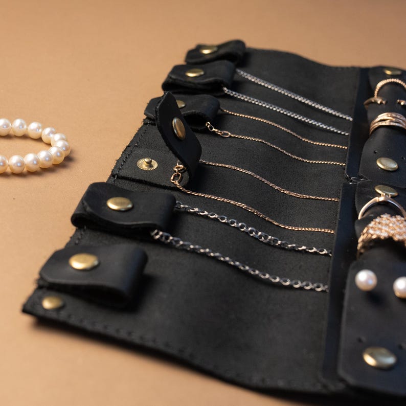 May include: A black leather jewelry roll organizer featuring multiple snap-button tabs to secure thin metal chains and a vertical strip with snap closures for rings and earrings, accompanied by a separate white pearl beaded bracelet.