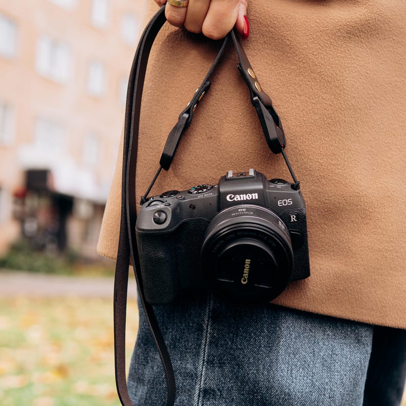 Puede incluir: Una c&aacute;mara Canon EOS R negra con una tapa de lente, colgada de una correa de cuero marr&oacute;n. La c&aacute;mara se sostiene contra un abrigo color canela y unos vaqueros azules. El logotipo de Canon es visible.