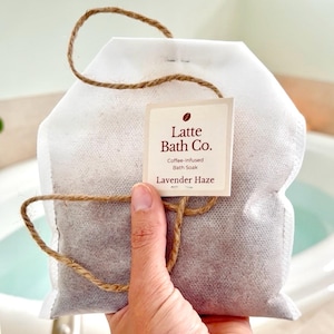 May include: A white sachet of coffee-infused bath soak labelled "Latte Bath Co. Lavender Haze." The sachet is held by a hand, with a natural twine loop and tag. The background shows a white bathtub.