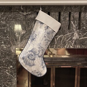May include: A white stocking with a blue and white seashell pattern hangs above a fireplace with a brass screen.