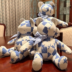 Può includere: Tre orsacchiotti di peluche realizzati in tessuto patchwork con un motivo ripetuto di stelle e forme geometriche blu e bianche. Ogni orso è decorato con un nastro a quadretti blu e bianco annodato attorno al collo.