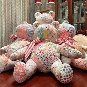 May include: A group of handmade patchwork teddy bears in various sizes, arranged on a dark wood table. The bears feature a variety of floral and geometric patterned fabrics in pastel colors, with red and white striped ribbon bows.