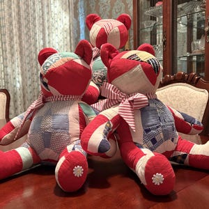 Puede incluir: Tres osos de peluche hechos de tela de retazos en tonos rojo, blanco y azul. Cada oso tiene una cinta a rayas rojas y blancas atada al cuello y un pequeño botón blanco en forma de flor en la planta de cada pie.
