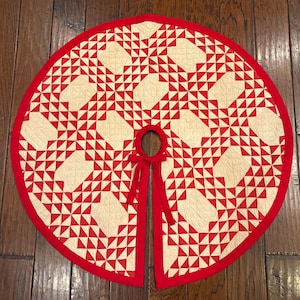 May include: A circular tree skirt featuring a red and cream-colored geometric quilt pattern. The fabric has a quilted texture with a solid red border and a center opening with two red fabric ties for closure, creating a symmetrical design.