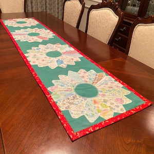 May include: A green table runner with a red border and a floral design. The runner features four large, white flowers with a green center, each made from smaller fabric pieces. The runner is on a wooden table.