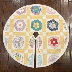 May include: A circular quilted tree skirt featuring a patchwork design of yellow and white squares. The quilt is decorated with various floral-shaped fabric appliqués in multiple colors and patterns, finished with a dotted fabric trim and ties.