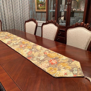 Puede incluir: Un camino de mesa marrón y beige con un patrón floral con flores rojas, amarillas y blancas. El camino de mesa está colocado sobre una mesa de madera oscura con sillas.