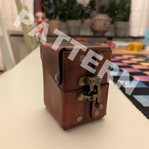 Op de afbeelding: Een bruine lederen doos in een vierkante vorm, voorzien van een scharnierend deksel dat wordt vastgezet met een messingkleurige metalen sluiting en decoratieve klinknagels op het voorpaneel. De doos heeft een gladde, matte afwerking en stiksels.