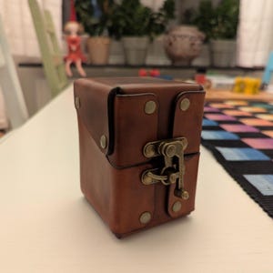 Op de afbeelding: Een rechthoekige doos van bruin leer met een scharnierend deksel, beveiligd met een messingkleurige metalen sluiting. De doos heeft zichtbare stiksels langs de randen en wordt bij elkaar gehouden door vier messingkleurige metalen klinknagels.