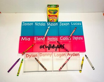 May include: A set of personalised name organisers in blue, pink, and clear plastic. Each organiser has a name label. Crayola crayons in various colours are scattered around the organisers and a box of 16 Crayola crayons is at the top.